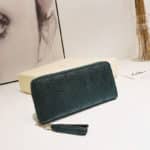 Pochette portefeuille vert eélégante avec des coutures apparentes. Pochette posé sur une table contre un support blanc devant une photo