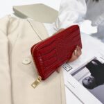 Portefeuille zippé long en cuir verni rouge crocodile tenu par une main de femme
