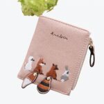 Portefeuille rose pâle avec des animaux cousues sur le tissu et leur queue qui dépasse du bord. Fond en bois marron et fleurs vertes sur le dessus de l'image