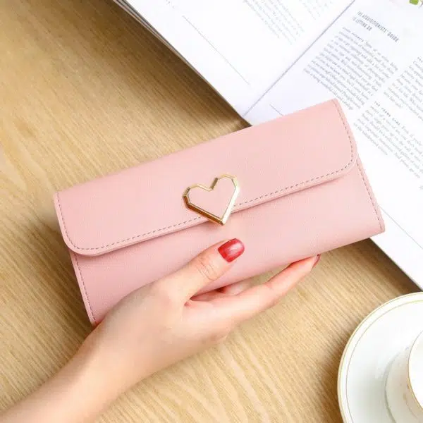 Portefeuille rectangulaire rose pâle tenu dans une main avec du vernis rouge. La main est posée sur une table en bois. Sur la table se trouve un journal ouvert et une tasse blanche avec sa soucoupe.