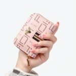Main de femme tenant un petit portefeuille rose avec des motifs sur fond gris.