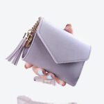 petit portefeuille violet clair en forme d'enveloppe avec un pompon. LE portefeuille et tenue dans un main devant un pull rose