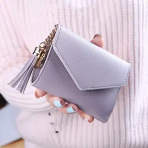 petit portefeuille violet clair en forme d'enveloppe avec un pompon. LE portefeuille et tenue dans un main devant un pull rose