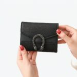 Portefeuille en cuir synthétiue noir tenue dans des mains avec du vernis rouge. Le portefeuille a une bboucle ne metal en forme de dragon