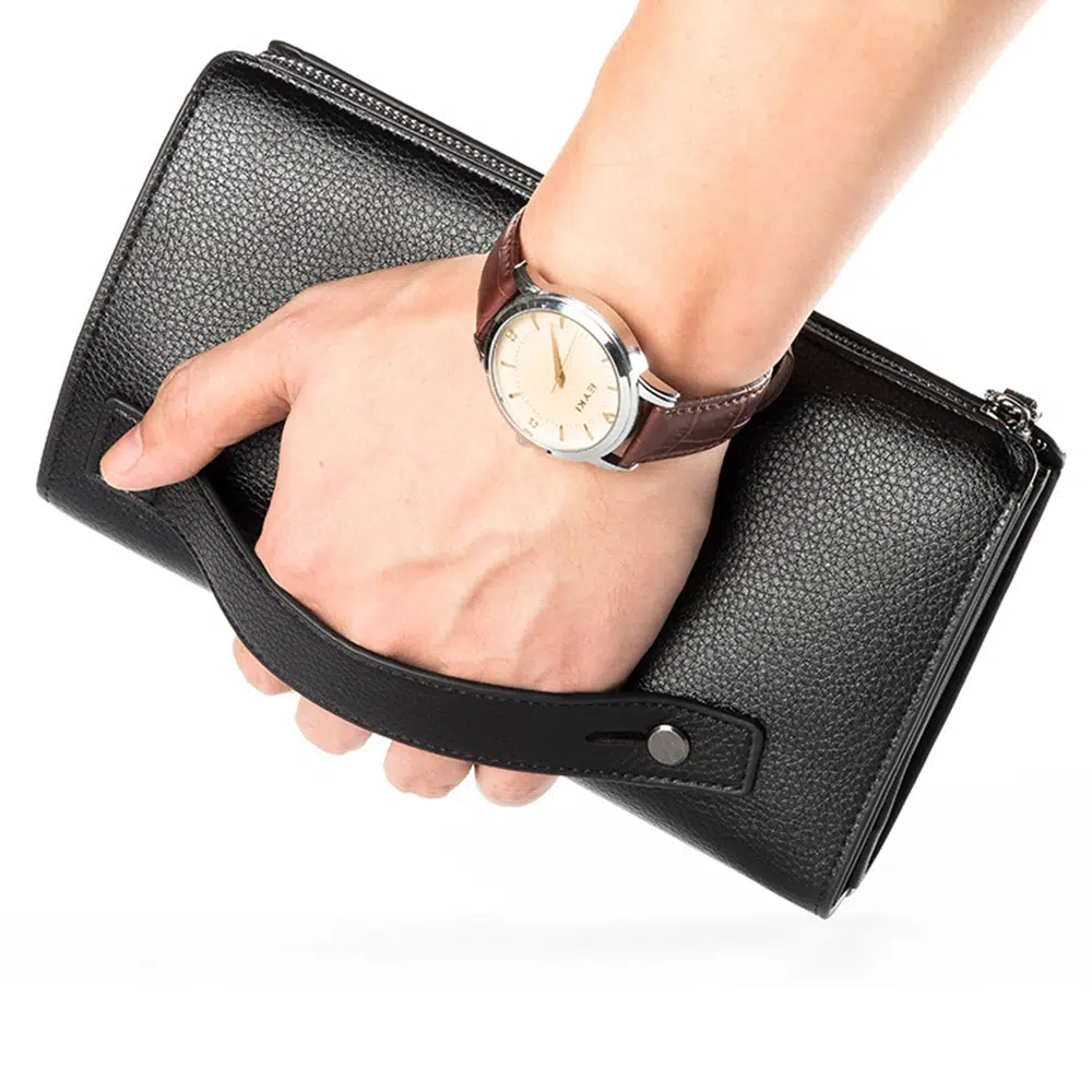 Main avec une montre tenant un portefeuille noir grâce à une anse. Fond blanc.