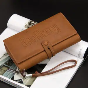 Pochette portefeuille marron clair avec dragonne et fermeture pression. Elle est posée sur un livre et il y a un fond noir.