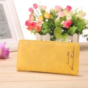 Joli chéquier portefeuille jaune de 18x9 cm, posé sur une table près d'un cadre et d'un bouquet de fleurs.