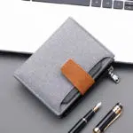Portefeuille en toile grise avec fermeture en cuir synthétique marron. Au fond, on voit un clavier d'ordinateur et des stylos au premier plan.