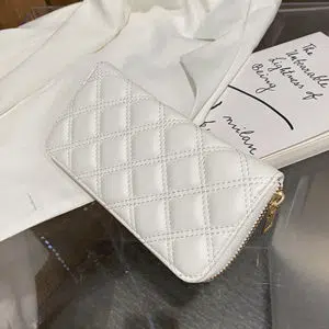 Pochette portefeuille blanc en cuir synthétique matelassé. LA pochette est posé sur une table en verre sur une veste blanche et un livrE.