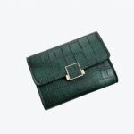 Portefeuille vert en crocodile posé sur une chemise blanche avec derrière un parquet marron