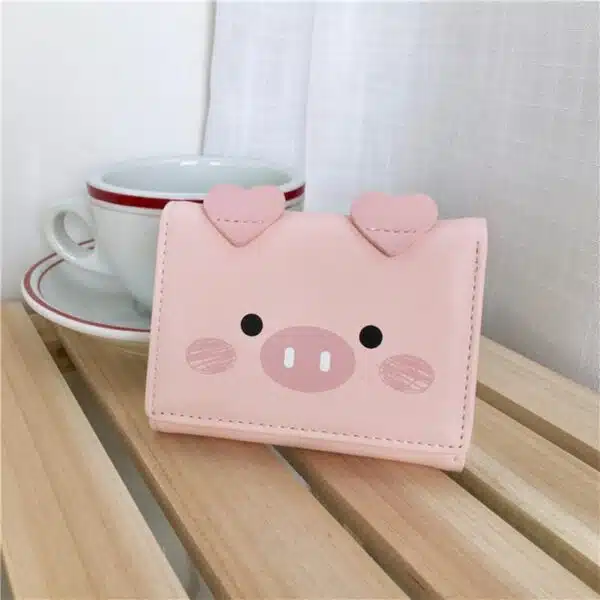 Portefeuille rose carré en forme de tête de cochon posé sur des lattes en bois et devant une tasse blanche dans sa soucoupe.