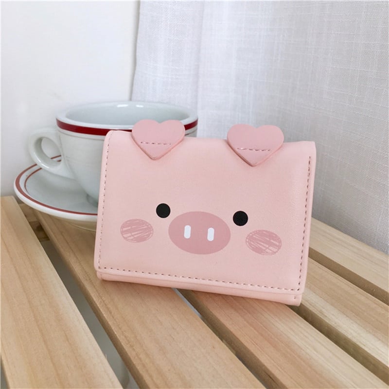 Portefeuille rose carré en forme de tête de cochon posé sur des lattes en bois et devant une tasse blanche dans sa soucoupe.